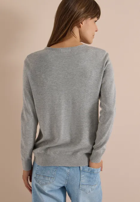 Frau von hinten, trägt grauen Pullover und blaue Jeans vor beigem Hintergrund.