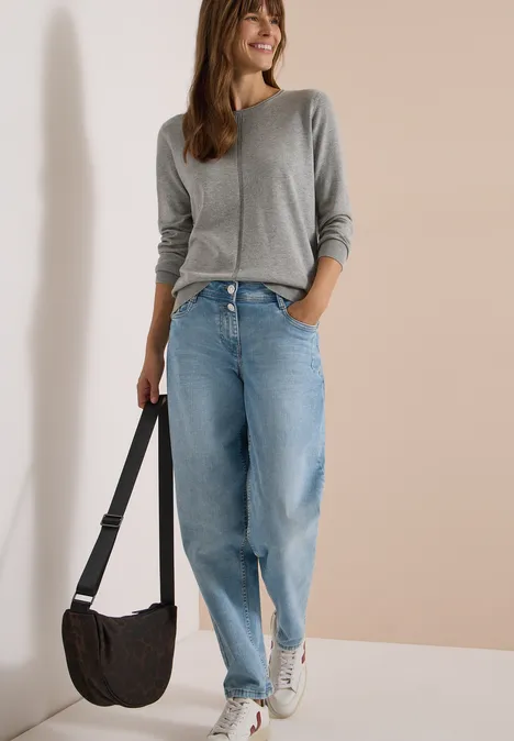 Junge Frau in grauem Pullover, weiten Jeans und Sneakers, mit Umhängetasche vor hellem Hintergrund.