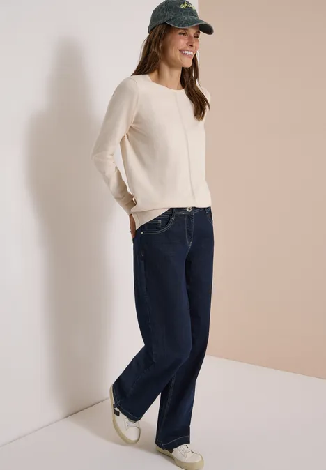 Basic Pullover mit Rundhals in Unifarbe oat milk beige melange Frau in beigem Pullover, breiten Jeans, weißen Sneakern und grüner Baseballkappe lehnt lächelnd in der Ecke.