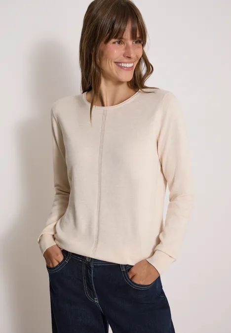 Basic Pullover mit Rundhals in Unifarbe oat milk beige melange Eine lächelnde Frau trägt einen hellbeigen Pullover und blaue Jeans vor hellem Hintergrund.