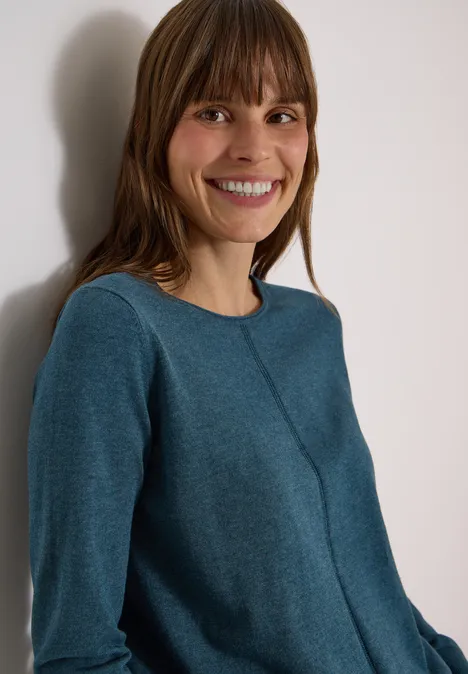 Basic Pullover mit Rundhals in Unifarbe dull teal blue melange Eine lächelnde Frau mit braunen Haaren in petrolfarbenem Pullover lehnt an einer weißen Wand.