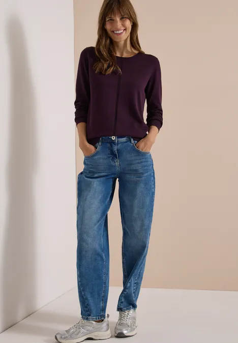 Basic Pullover mit Rundhals in Unifarbe eggplant red Frau lehnt entspannt an einer Wand, trägt lila Pullover, blaue Jeans und silberne Sneakers.