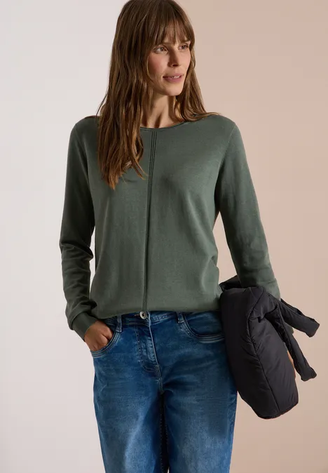 Basic Pullover mit Rundhals in Unifarbe dried olive green Frau in grünem Pullover, Jeans und hält eine dunkle Jacke in der Hand.