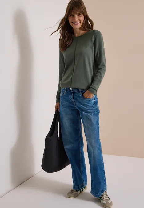 Basic Pullover mit Rundhals in Unifarbe dried olive green Frau in legerem Outfit: grünes Langarmshirt, lockere Jeans, Sneaker und schwarze Tasche vor weißer Wand.