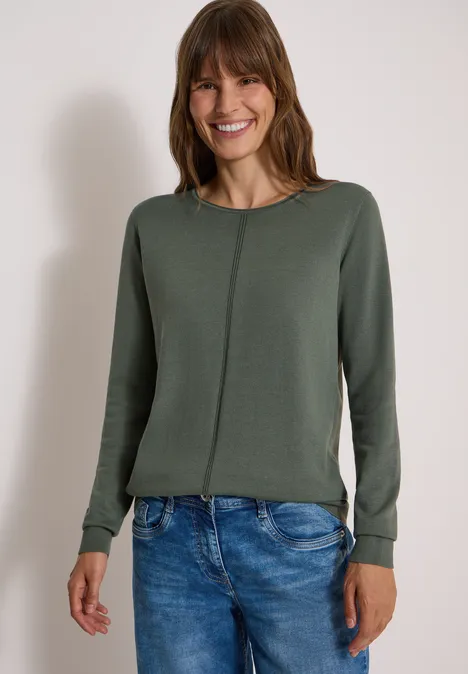 Basic Pullover mit Rundhals in Unifarbe dried olive green Eine lächelnde Frau in grünem Pullover und blauen Jeans vor hellem Hintergrund.