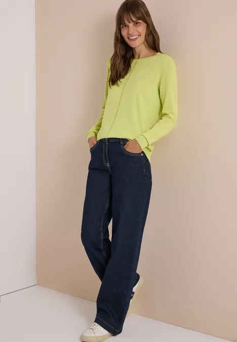 Basic Pullover mit Rundhals in Unifarbe citrin green Frau in limettengrünem Pullover, weiten Jeans und weißen Sneakern lehnt lächelnd entspannt an der Wand.
