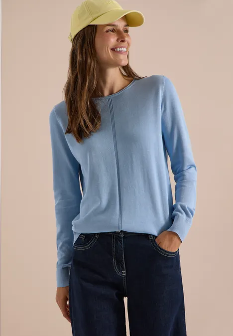 Basic Pullover mit Rundhals in Unifarbe easy blue Frau in hellblauem Pullover, Jeans und gelbgrüner Kappe vor neutralem Hintergrund, lächelnd seitlich schauend.