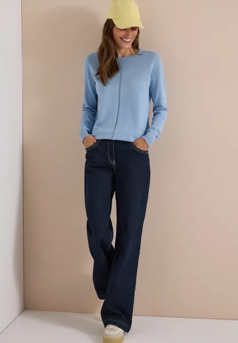 Basic Pullover mit Rundhals in Unifarbe easy blue Eine lächelnde Frau trägt eine gelbe Kappe, einen hellblauen Pullover, weite dunkle Jeans und weiße Sneaker.