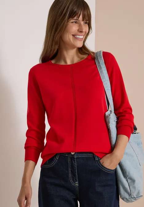 Basic Pullover mit Rundhals in Unifarbe flame red Eine lächelnde Frau in rotem Pullover und Jeans mit hellblauer Tasche über der Schulter.