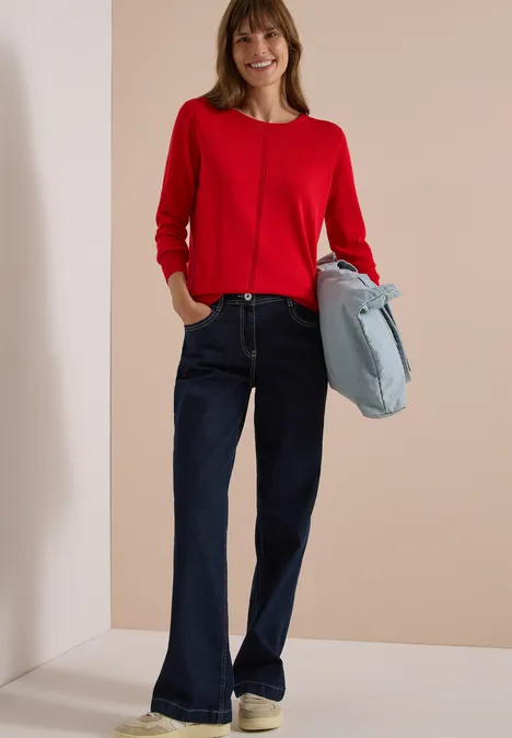 Basic Pullover mit Rundhals in Unifarbe flame red Eine Frau lächelt, trägt roten Pullover, dunkelblaue Schlagjeans und hält eine hellblaue Tasche.