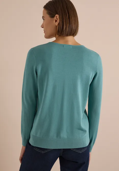 Pull basique uni à col rond mineral blue Une femme de dos porte un pull turquoise et un jean sur un fond neutre.