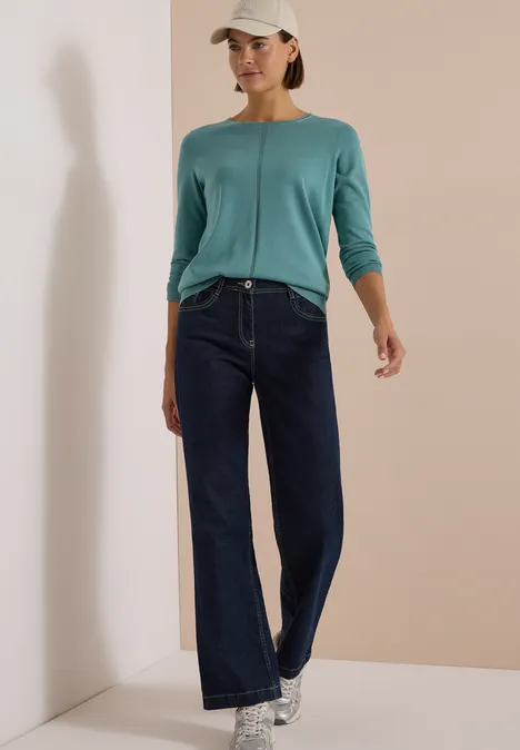 Pull basique uni à col rond mineral blue Femme portant un haut bleu-vert à manches longues, jean large, baskets et casquette devant un mur beige.