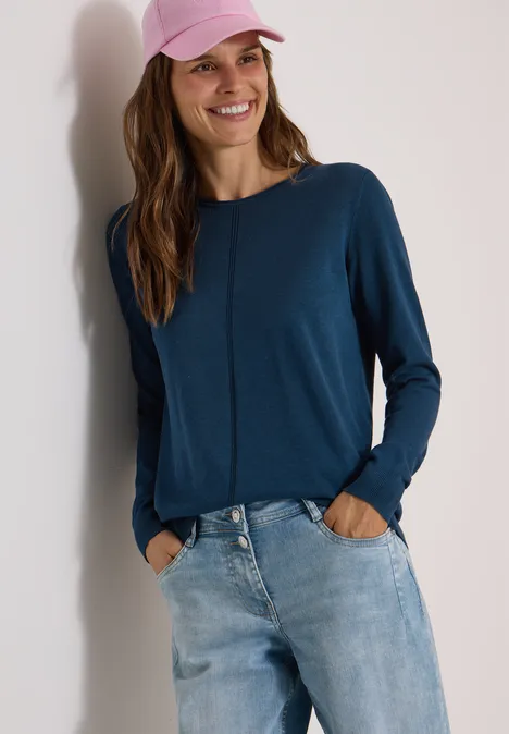 Basic Pullover mit Rundhals in Unifarbe moonlight blue Eine Frau mit blauem Langarmshirt, Jeans und rosa Cap lehnt an einer Wand und lächelt.