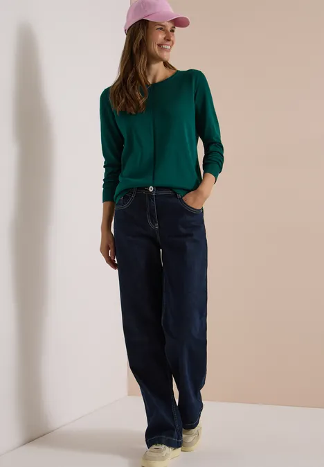 Basic Pullover mit Rundhals in Unifarbe dark fir green Eine Frau steht lässig in Lockermode: grüner Pullover, weite Jeans, beige Sneaker und rosa Baseballcap.