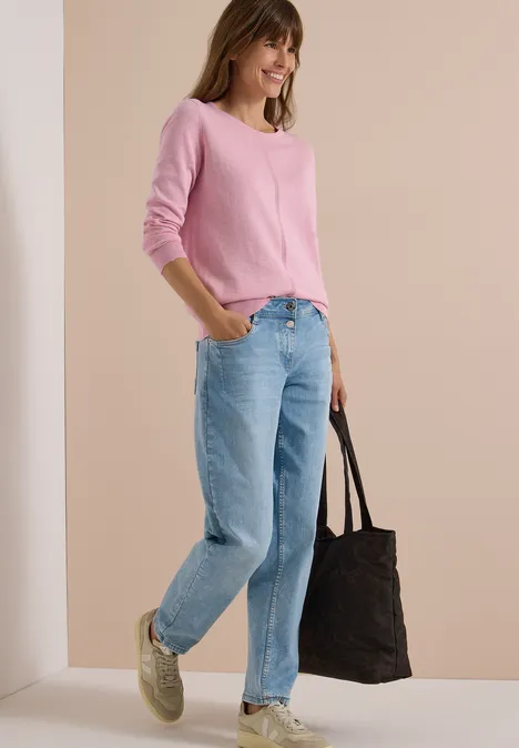 Frau in pinkem Pullover und Jeans, trägt grüne Sneakers und schwarzen Shopper, stehend vor hellem Hintergrund.