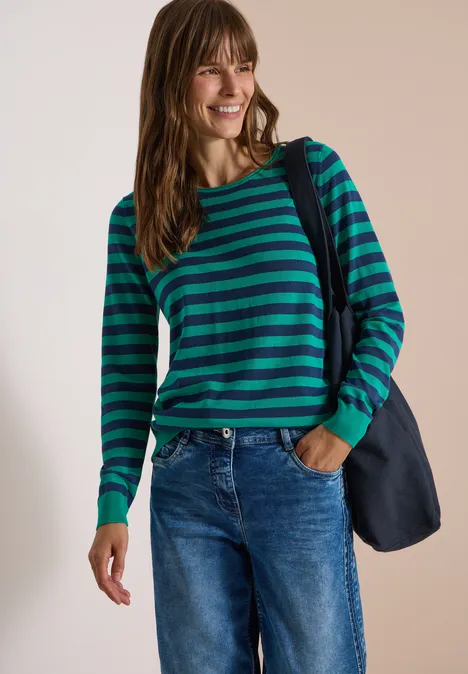 Frau in grün-blau gestreiftem Pullover und Jeans, lächelnd mit Umhängetasche vor neutralem Hintergrund.