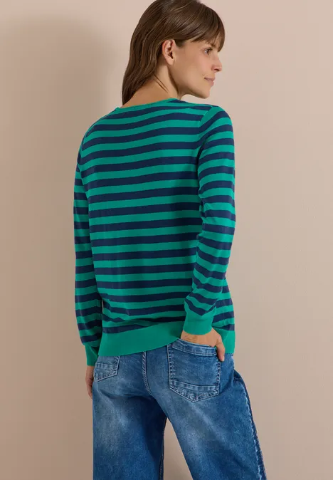 Rückenansicht einer Person in grün-blau gestreiftem Pullover und blauen Jeans vor beigem Hintergrund.
