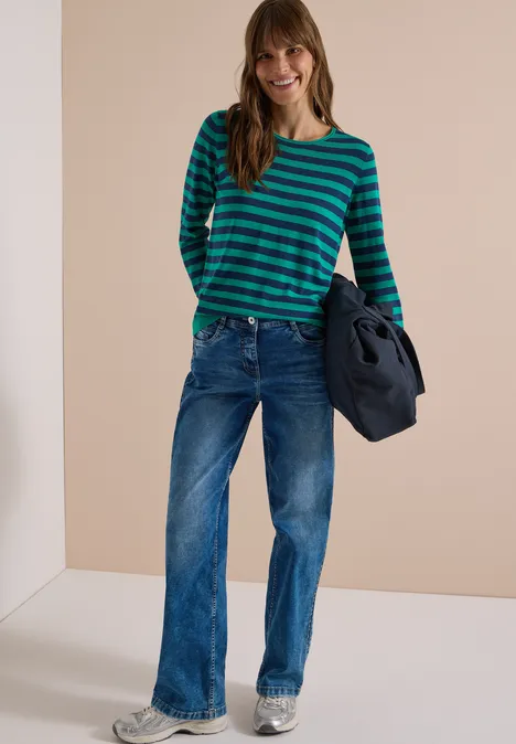 Frau in grün-blau gestreiftem Pullover und weiten Jeans, hält Jacke, lächelt vor einfachem Hintergrund.
