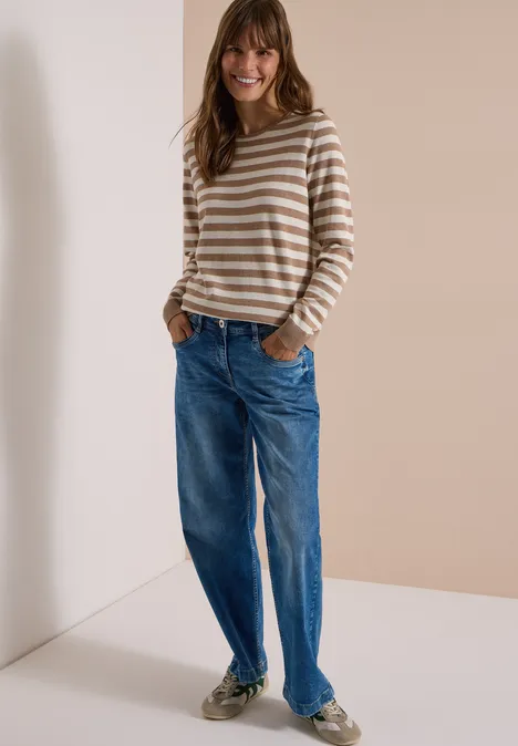 Frau mit gestreiftem Langarmshirt, blauen Jeans und Sneakern lehnt an heller Wand und lächelt.