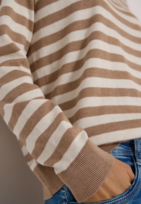 Ein Oberkörper in beigem, weiß gestreiftem Pullover und blauen Jeans mit einer Hand in der Tasche.