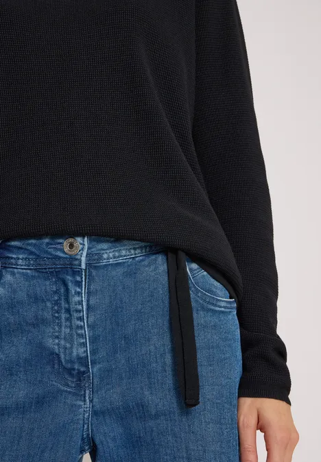 Person trägt schwarzen Pullover und blaue Jeans, nur Oberkörper bis Hüfte sichtbar.