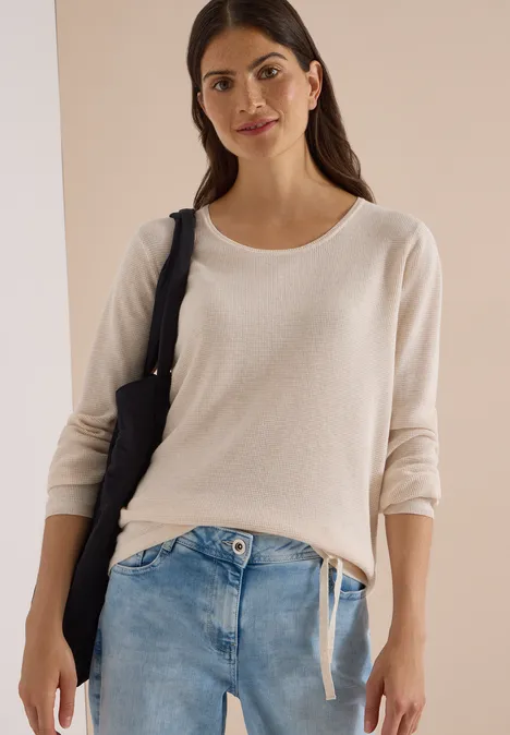 Frau in hellem Pullover mit 3/4-Ärmeln, blauer Jeans und dunkler Schultertasche, heller Hintergrund.