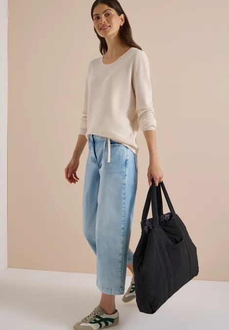 Frau in beigem Pullover, weiten Jeans und Sneakers trägt große, schwarze Stepp-Tasche.
