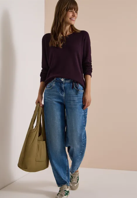 Pullover mit Rundhals und Struktur eggplant red Frau in lila Pullover, weiten Jeans und Sneakers, trägt eine große grüne Tasche und lächelt entspannt.