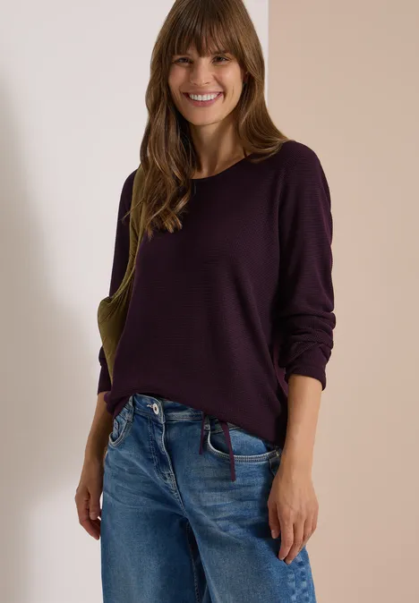 Pullover mit Rundhals und Struktur eggplant red Frau mit lila Pullover und Jeans lächelt vor neutralem Hintergrund.