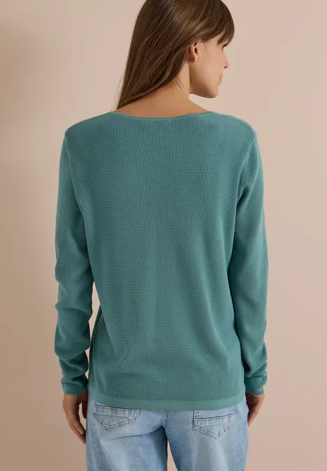 Eine Frau von hinten trägt einen türkisfarbenen Pullover und Jeans vor schlichtem Hintergrund.