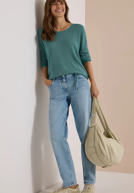 Eine Frau lehnt sich an eine Wand, trägt eine grüne Bluse, blaue Jeans und eine große Umhängetasche.