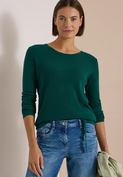 Pullover mit Rundhals und Struktur dark fir green Frau mit dunkelgrünem Pullover und blauen Jeans steht vor neutralem Hintergrund und hält eine Jacke.