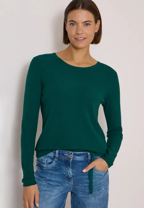 Pullover mit Rundhals und Struktur dark fir green Junge Frau mit dunkelgrünem Pullover und Jeans lächelt entspannt mit einer Hand in der Hosentasche.