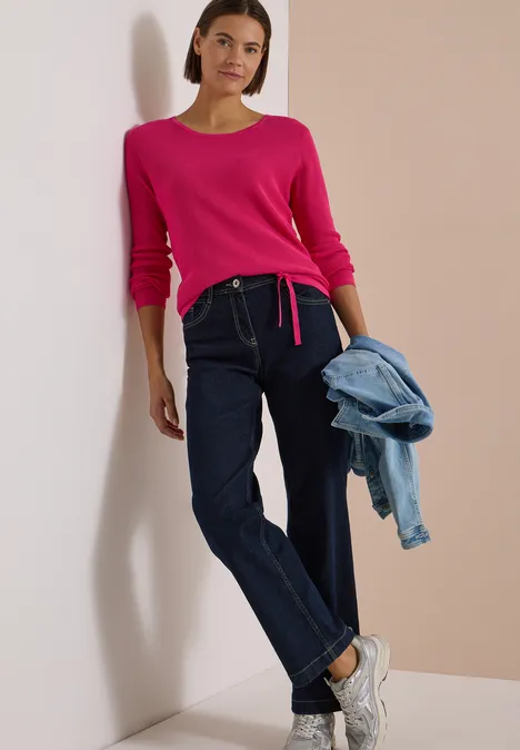 Pullover mit Rundhals und Struktur pink flash Frau lehnt an Wand in pinkem Pullover, dunklen Jeans, weißen Sneakers und hält eine Jeansjacke in der Hand.