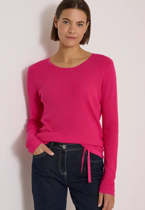 Pullover mit Rundhals und Struktur pink flash Frau in pinkem Rundhals-Pullover mit seitlichem Bindeband und dunkler Jeans vor hellem Hintergrund.