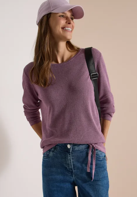 Pullover mit Rundhals und Struktur lavender purple melange Junge Frau lächelt, trägt violettes Shirt, Jeans, Cap und Umhängetasche.