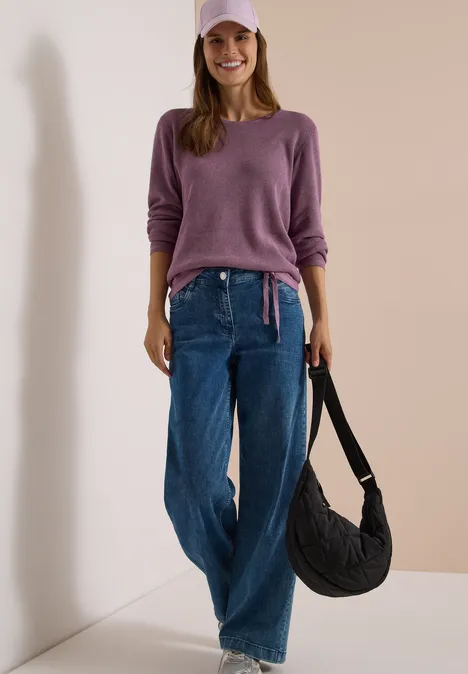Pullover mit Rundhals und Struktur lavender purple melange Eine lächelnde Frau in lila Pullover, passender Cap, weiten Jeans und schwarzer Schultertasche lehnt an einer Wand.