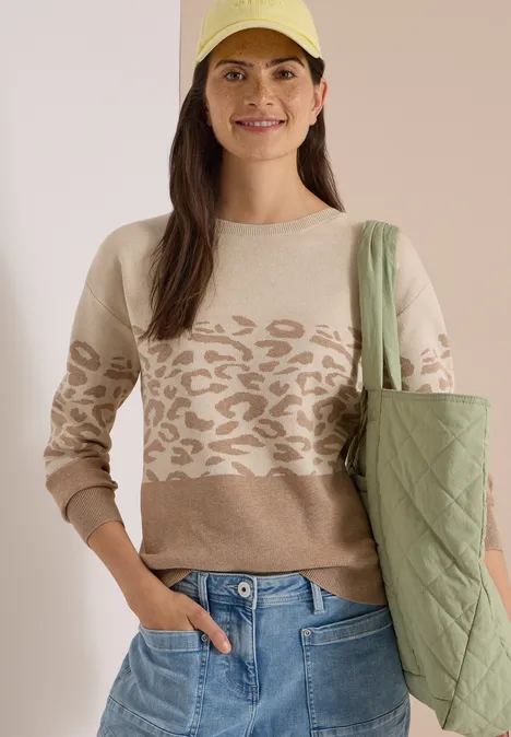 Langarm Pullover mit Leo-Jacquard-Muster creamy mocha melange Frau in beigem Pullover mit Leo-Muster, blauen Jeans, grüner gesteppter Tasche und gelbem Stirnband.