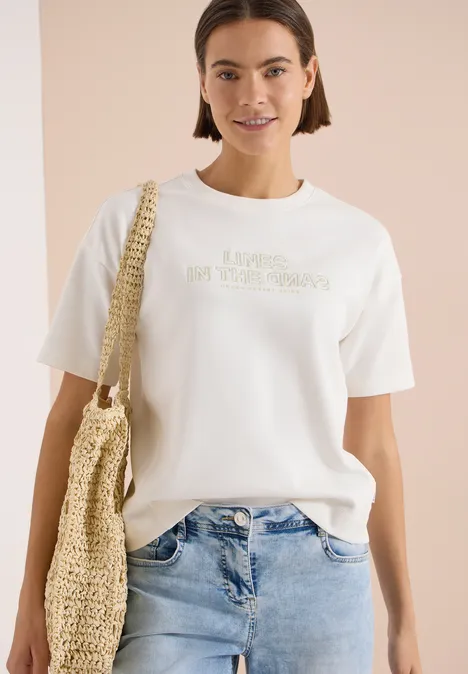 Vrouw in wit t-shirt, spijkerbroek en rieten tas voor een neutrale achtergrond.