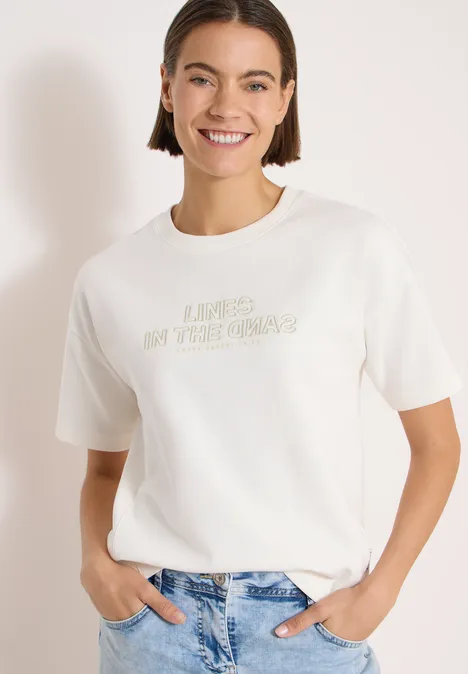 Vrouw draagt wit t-shirt met opdruk en blauwe spijkerbroek tegen witte achtergrond.