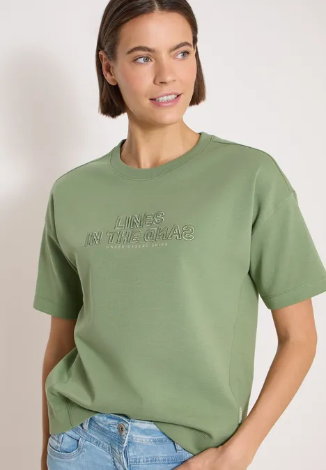Persoon draagt olijfgroen t-shirt met print en een lichtblauwe spijkerbroek