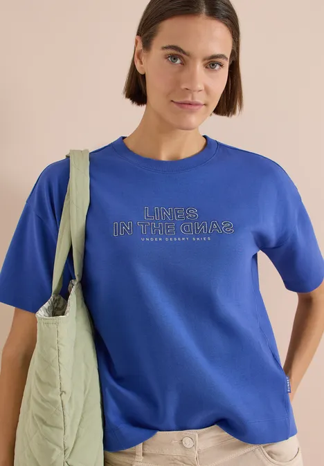 Vrouw in blauw T-shirt met tekst 'Living in the chaos' en groene gewatteerde schoudertas.