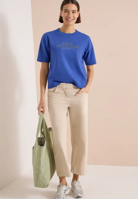 Vrouw met blauw T-shirt, beige broek en groene boodschappentas voor neutrale achtergrond.
