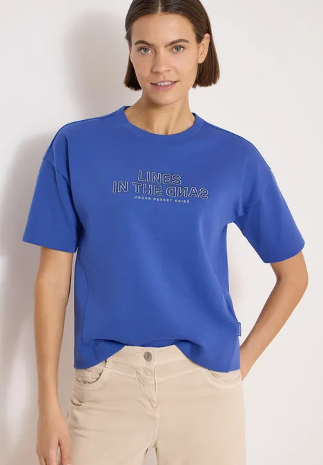 Persoon voor een lichte achtergrond draagt een blauw T-shirt met tekst en een lichte broek.