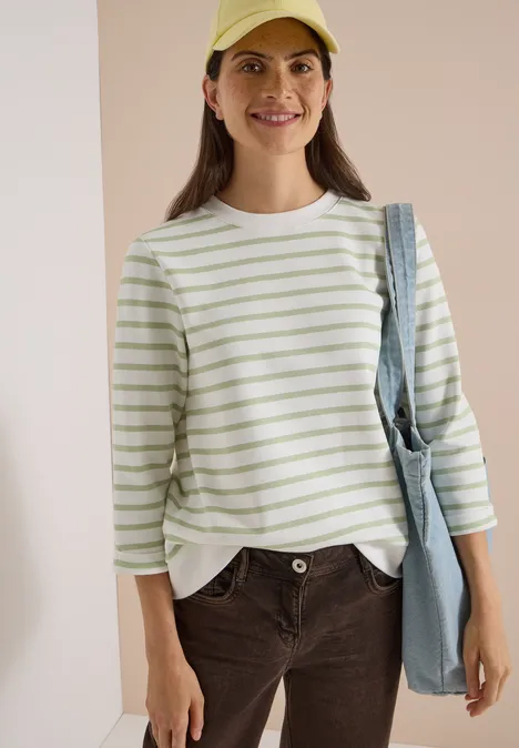 Een vrouw glimlacht voor een lichte achtergrond, terwijl ze een gestreept shirt, pet en tas draagt.