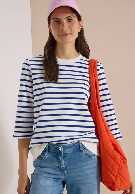 3/4-Arm Sweatshirt mit Rundhals und Streifen deep river blue Frau mit gestreiftem Shirt, Jeans, roter Kappe und orangefarbener Tasche vor neutralem Hintergrund.