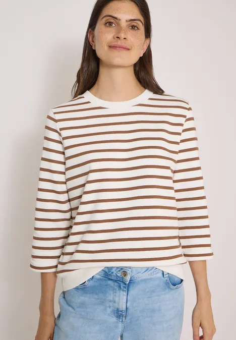 Vrouw in wit-bruin gestreepte trui en blauwe jeans, vooraanzicht, los haar, subtiele glimlach.