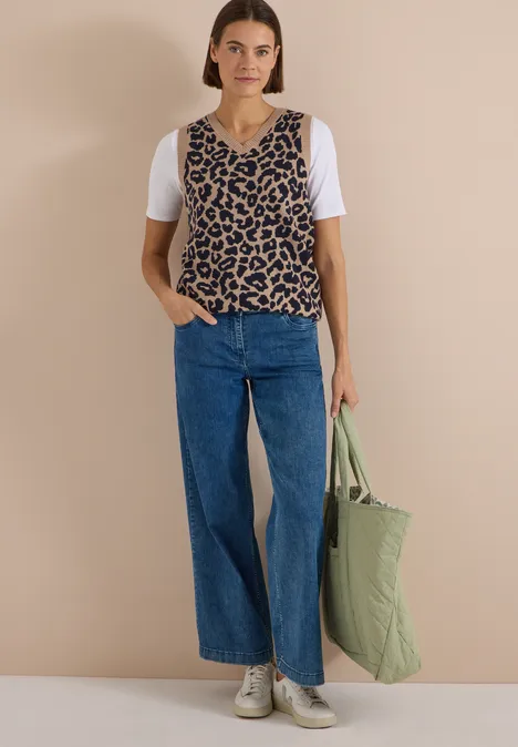Frau mit weißem T-Shirt, Leo-Top, Jeans, weißen Sneakern und grünem Stepp-Shopper vor beigem Hintergrund.