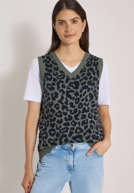 Frau trägt grüne Strickweste mit schwarzem Leopardenmuster, weißem T-Shirt und blauen Jeans.