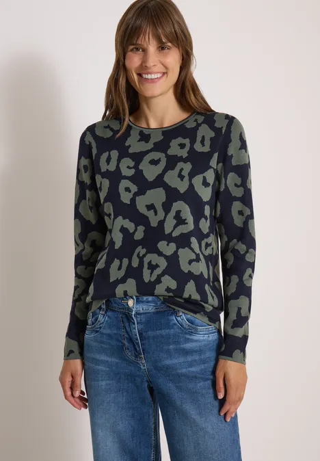Frau in lässigem Animal-Print-Pullover und blauen Jeans, lächelt vor hellem Hintergrund.